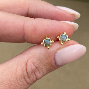 May include: A pair of gold stud earrings with small square labradorite stones. The stones have a dark grey and blue colour.