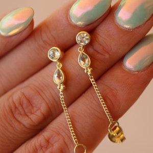 May include: A pair of gold dangle earrings with a teardrop-shaped blue gemstone and a round blue gemstone. The earrings have a delicate gold chain that hangs down from the gemstones.