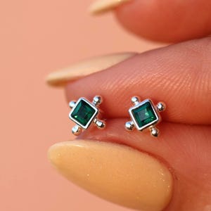 Könnte beinhalten: Ein Paar kleiner Ohrstecker mit quadratischen, smaragdgrünen Edelsteinen. Die Ohrringe sind in silberfarbenem Metall mit einem Vier-Punkt-Design gefasst, wobei jeder Punkt einen kleinen, runden Akzent aufweist. Auf weichem Pfirsichhintergrund.