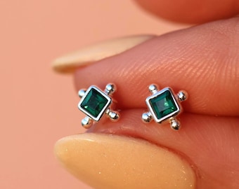 Puede incluir: Un par de pendientes pequeños con piedras preciosas cuadradas de color verde esmeralda. Los pendientes están engastados en metal plateado con un diseño de cuatro puntas, cada punta con un pequeño acento redondo. Fondo melocotón.