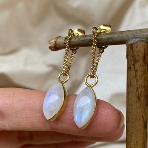 Puede incluir: Un par de pendientes de oro con una cadena delicada y un colgante de piedra lunar nacarada. La piedra lunar es de un blanco claro e iridiscente con un brillo sutil.