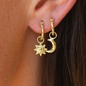 Puede incluir: Aretes de aro dorados con un pequeño dije de sol y luna colgando de la parte inferior.