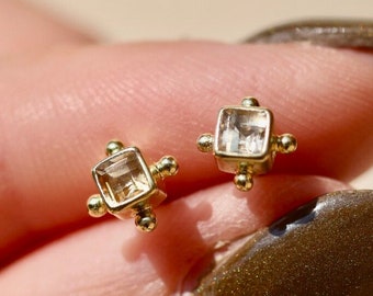 May include: A pair of small, square-shaped stud earrings. Each earring features a clear, square-cut gemstone set in a gold-colored metal setting. The earrings are held between fingers, showcasing their delicate design.