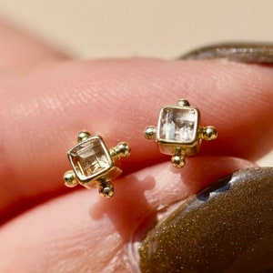 Puede incluir: Un par de pendientes pequeños y cuadrados. Cada pendiente presenta una piedra preciosa transparente de corte cuadrado engastada en un engaste de metal dorado. Los pendientes se sostienen entre los dedos.