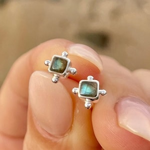 May include: A pair of silver stud earrings featuring square-shaped labradorite gemstones. The earrings have a simple design with small silver beads around the edges of the gemstones.