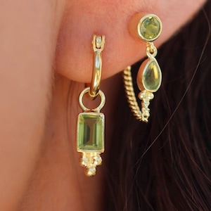 May include: A pair of gold earrings with green gemstones. The earrings are a hoop with a dangling rectangular stone and a smaller stud with a round stone.