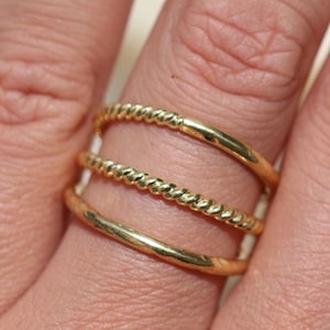 Puede incluir: Un anillo de oro con tres bandas. La banda superior es un diseño de cuerda trenzada, la banda del medio es una banda lisa y la banda inferior es una banda lisa.