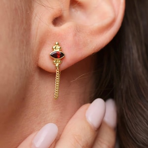 May include: Gold-toned earring with a dangling chain. The earring features a red, oval-shaped stone at the top, with a chain that hangs down. The earring is attached to the ear. Jewelry.