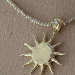 May include: Gold-toned sunburst pendant necklace with a delicate pearl strand. The sun pendant has a textured surface and pointed rays. A small, round, iridescent stone is set in the bail. The necklace is on a neutral background.
