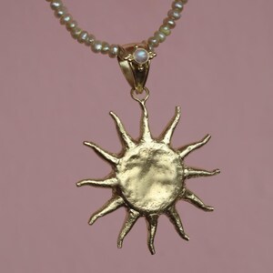 May include: A gold-colored sun pendant necklace with a pearl beaded chain. The sun pendant has a textured surface and pointed rays. A small pearl is set in the gold clasp. The background is a solid pink color.