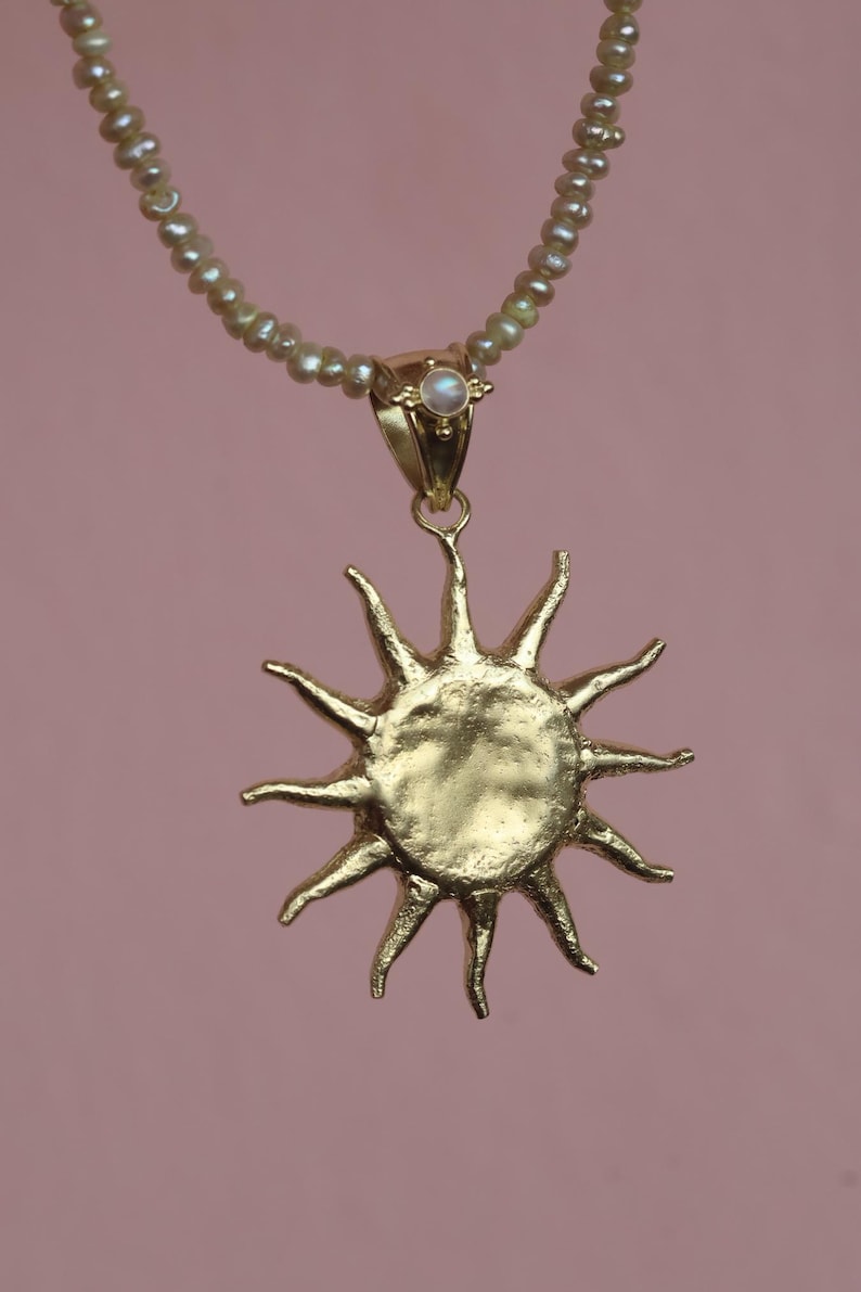 May include: A gold-colored sun pendant necklace with a pearl beaded chain. The sun pendant has a textured surface and pointed rays. A small pearl is set in the gold clasp. The background is a solid pink color.