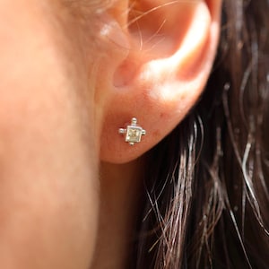 May include: A close-up of a silver stud earring with a square, light-colored gemstone. The earring is set in a square frame with small, round accents at each corner. The earring is in the ear of a person.