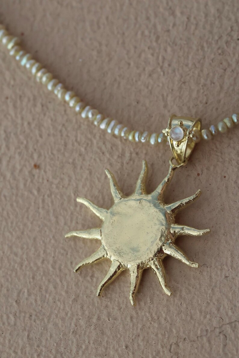 May include: Gold-toned sunburst pendant necklace with a delicate pearl strand. The sun pendant has a textured surface and pointed rays. A small, round, iridescent stone is set in the bail. The necklace is on a neutral background.
