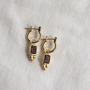 May include: A pair of gold-coloured hoop earrings. Each earring has a rectangular, dark-coloured gemstone pendant. The pendant hangs from a small hoop and has a cluster of three small, round accents at the bottom.