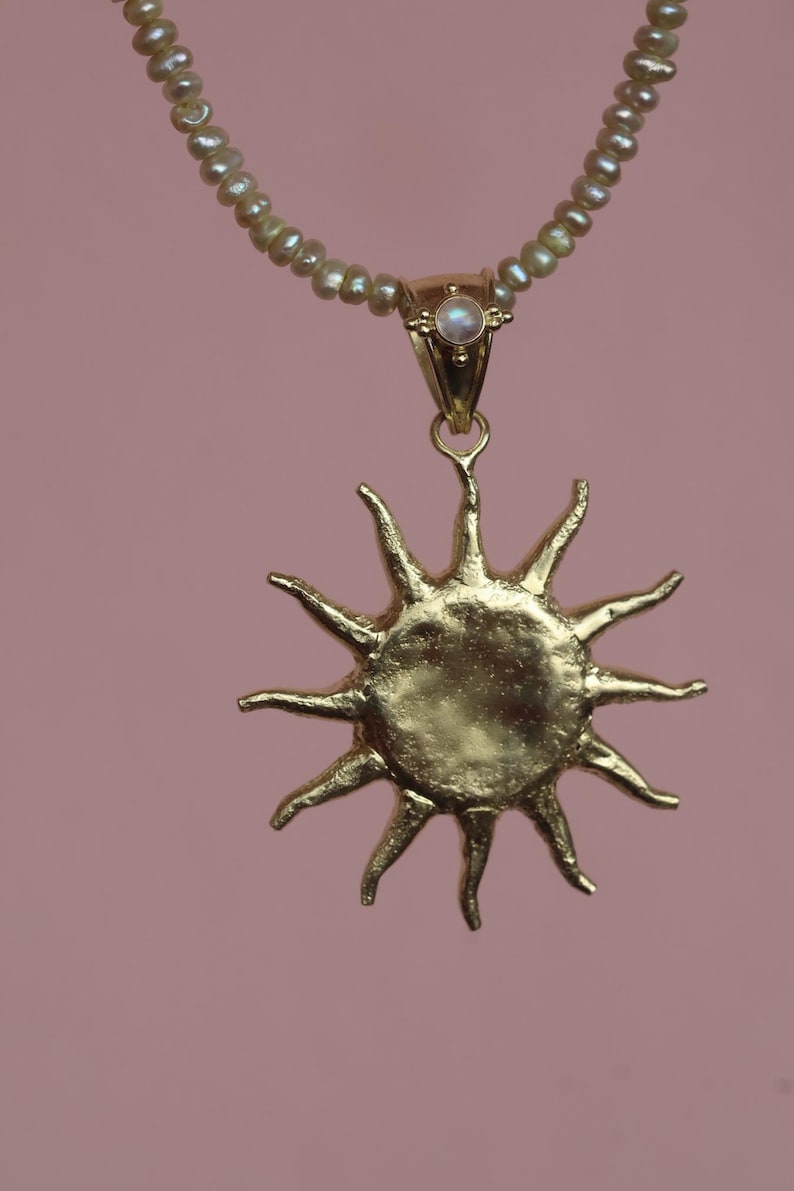May include: A gold-colored sun pendant necklace with a pearl beaded chain. The sun pendant has multiple rays extending outward. A small, round, iridescent stone is set in the gold clasp. The background is a solid, muted pink.