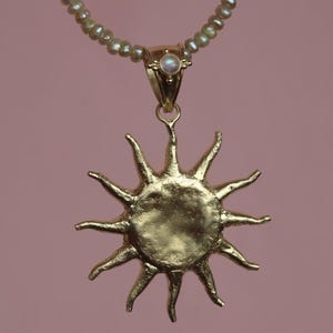 May include: A gold-colored sun pendant necklace with a pearl beaded chain. The sun pendant has a textured surface and pointed rays. A small, round, iridescent stone is set in the pendant's bail. The background is a solid, muted pink.