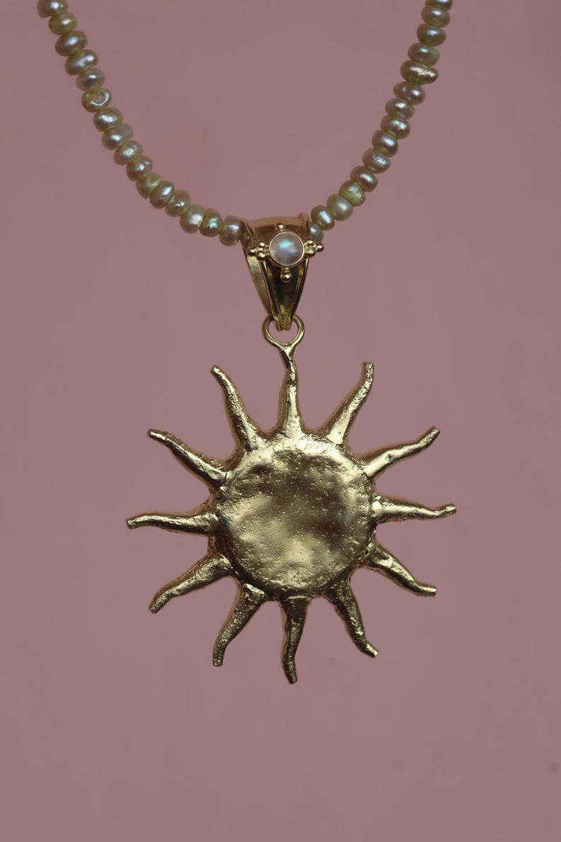 May include: A gold-colored sun pendant necklace with a pearl beaded chain. The sun pendant has a textured surface and pointed rays. A small, round, iridescent stone is set in the pendant's bail. The background is a solid, muted pink.