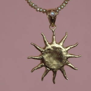 May include: A gold-colored sun pendant necklace with a pearl beaded chain. The sun pendant has multiple rays extending outward. A small, round, iridescent stone is set in the gold clasp. The background is a solid, muted pink.