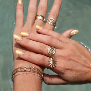 May include: Close-up of hands with yellow nail polish wearing gold rings. The rings feature a sun, a spiral, and an evil eye design. The hands are wearing a gold bracelet with a moon and star design.