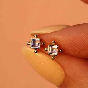 Pode incluir: Um par de brincos de botão dourados. Cada brinco apresenta uma pedra preciosa quadrada, roxa clara, emoldurada em ouro. Pequenas contas douradas são colocadas em cada canto do quadrado.
