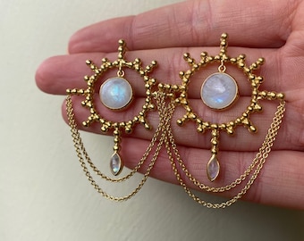 Può includere: Orecchini dorati con un ciondolo circolare in pietra di luna e catene pendenti. Gli orecchini hanno un design a forma di sole con un bordo di perline.