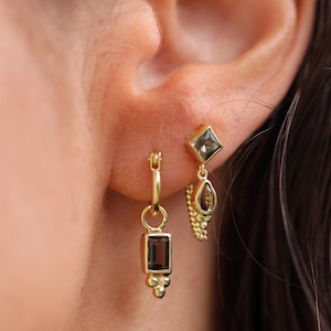 May include: Close-up of two gold earrings. One is a hoop with a rectangular brown stone and small gold beads. The other is a square-shaped stone with a teardrop-shaped stone hanging below it, connected by a gold chain.