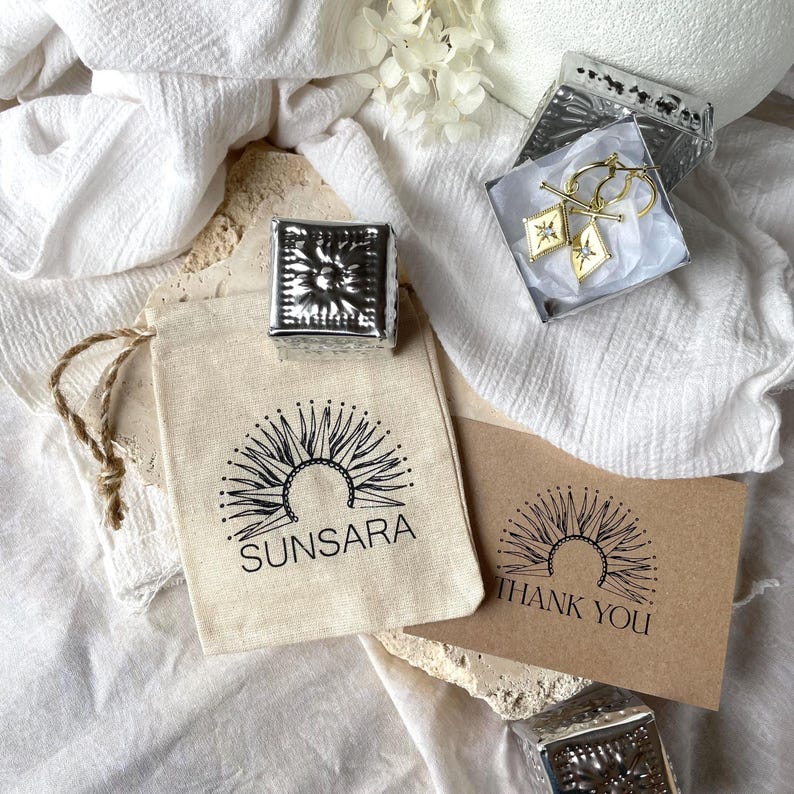 May include: An overhead shot showcases jewelry and packaging. A small silver box with a sunburst design sits atop a canvas bag printed with "SUNSARA." Gold hoop earrings with diamond-shaped charms are in a box. A thank you card is also visible.