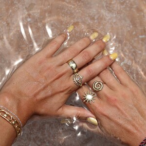 May include: Close-up of hands wearing multiple gold rings, including a sun ring, an eye ring, and a simple band. The hands are submerged in clear water with a light brown background.