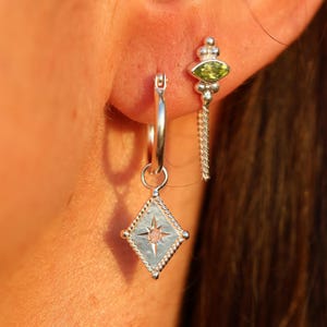 Peut inclure: Boucle d'oreille argentée avec un pendentif en forme de losange orné d'un motif en étoile et d'une petite pierre rose. Une deuxième boucle d'oreille présente une pierre verte et une chaîne. Bijoux.