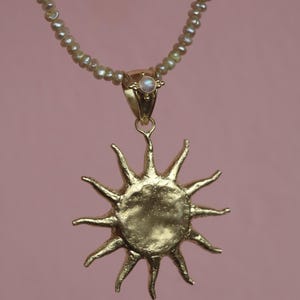 May include: A gold-colored sun pendant necklace with a pearl beaded chain. The sun pendant has a textured surface and pointed rays. The necklace has a small pearl at the top of the pendant and a pink background.