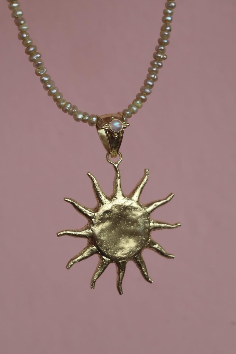 May include: A gold-colored sun pendant necklace with a pearl beaded chain. The sun pendant has a textured surface and pointed rays. The necklace has a small pearl at the top of the pendant and a pink background.