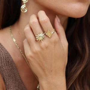 May include: A woman's hand wearing two gold rings. One ring features a sun design, and the other ring has a geometric diamond shape.