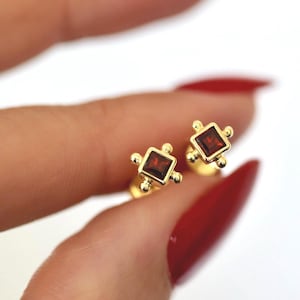 May include: A pair of gold earrings with small square red gemstones. The earrings have a geometric design with small gold balls around the gemstones.