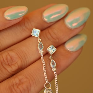 May include: A pair of silver dangle earrings with blue gemstone accents. The earrings feature a square-shaped gemstone at the top and a teardrop-shaped gemstone below. The earrings are suspended from a delicate silver chain.