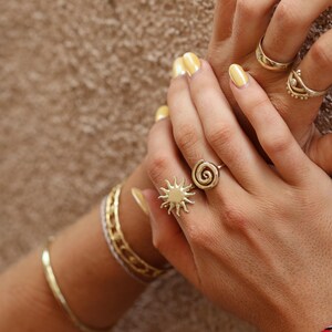 May include: A person's hands wearing multiple gold rings, including a sun and spiral design ring. The person is also wearing several bracelets on their wrist.