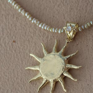 May include: Gold-toned sunburst pendant necklace with a delicate pearl strand. The sun pendant has a textured surface and pointed rays. A small, round, iridescent stone is set in the bail. The necklace is on a neutral background.