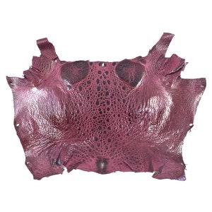 May include: A piece of dark pink ostrich leather with a textured surface. The leather is cut into a rectangular shape with rounded corners.
