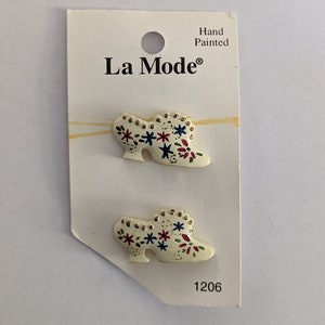 May include: Two white hand-painted wooden buttons shaped like shoes with floral designs in red, blue, and gold. The buttons are on a white card with the text "La Mode®" and "Hand Painted" and the number "1206".