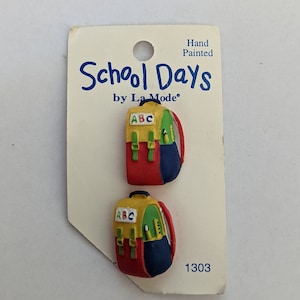 May include: Two hand-painted plastic buttons shaped like school backpacks. The backpacks are yellow, red, blue, and green with the letters "ABC" on the front. The buttons are labeled "School Days by La Mode" and "1303".