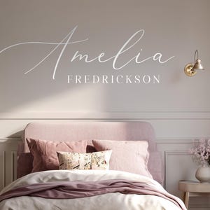 May include: A bedroom interior with a pink bed, cushions, and bedding. The wall above the bed features the name "Amelia Fredrickson" in a stylish script. The room is decorated in neutral tones, with wall sconces and a patterned rug adding detail.