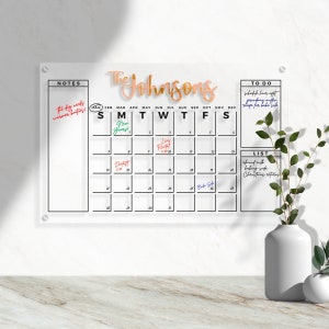 May include: A white dry-erase calendar with a black frame and a black grid. The calendar has the months of the year listed across the top and the days of the week listed down the side. The calendar is labelled "The Johnsons" in gold lettering. There are notes written on the calendar, including "The dog needs warmer booties!" and "New Years!" There is a to-do list on the right side of the calendar with the following items: "Schedule hair appt", "Grandmother muffin", "Recipe for bake sale". There is also a list labelled "List" with the following items: "Almond milk", "Baking soda", "Christmas ribbons".