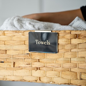May include: A wicker basket with a black label that says "Towels" on it. The basket is filled with white towels.