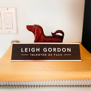 A wooden dog figurine is sitting on a yellow surface, with a brown sign that reads "LEIGH GORDON TALENTED AS FUCK" in front of it.