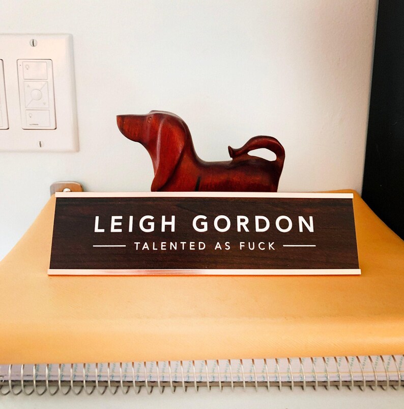 A wooden dog figurine is sitting on a yellow surface, with a brown sign that reads "LEIGH GORDON TALENTED AS FUCK" in front of it.
