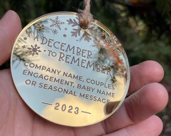 以下が含まれることがあります： 雪の結晶が刻印された円形のゴールドミラーオーナメント。A DECEMBER TO REMEMBER, COMPANY NAME, COUPLES ENGAGEMENT, BABY NAME OR SEASONAL MESSAGE, 2023という文字が刻まれています。茶色の麻紐のループで吊り下げられています。