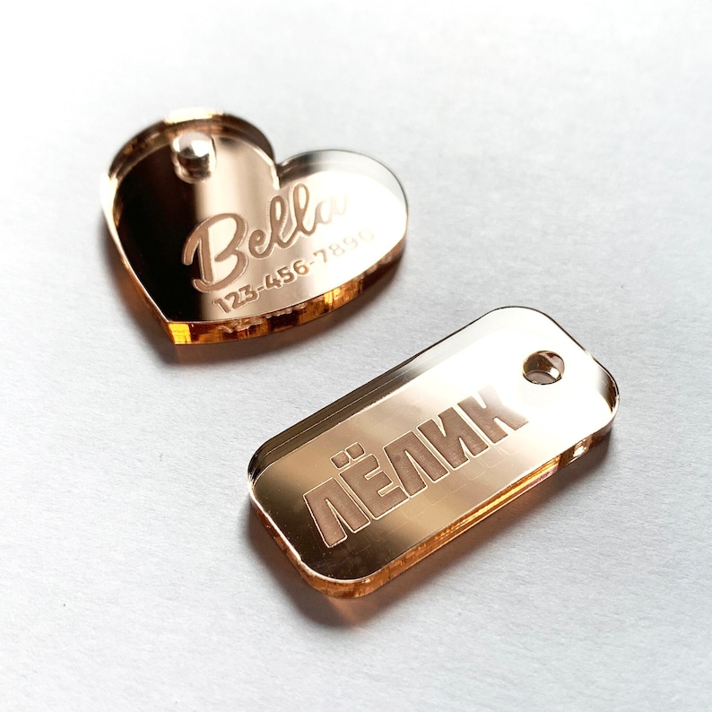 May include: Two rose gold pet tags. One is heart-shaped with the name "Bella" and a phone number. The other is rectangular with Cyrillic text. Both tags have a hole for attachment.
