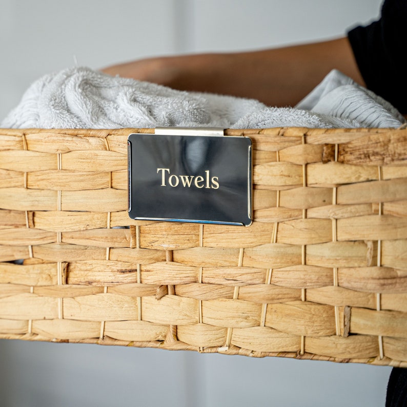 Puede incluir: Una cesta de mimbre con una etiqueta negra que dice "Towels". La cesta est&aacute; llena de toallas blancas.