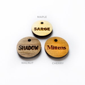 May include: Three round wooden tags with laser-etched text. The top tag is maple with "Sarge", the middle tag is walnut with "Shadow", and the bottom tag is cherry with "Mittens". Each tag has a small hole at the top.