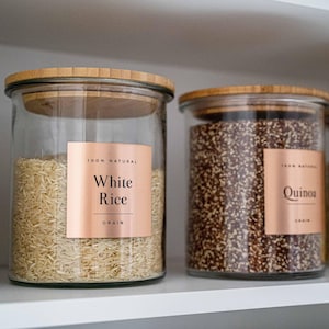 Könnte beinhalten: Zwei Vorratsgläser aus klarem Glas mit hellbraunen Bambusdeckeln. Das linke Glas enthält weißen Reis und hat ein kupferfarbenes Etikett mit dem Text 100% NATURAL White Rice GRAIN. Das rechte Glas enthält Quinoa mit einem passenden Etikett.