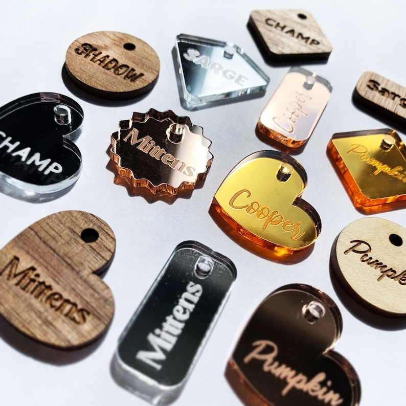 May include: A collection of various pet tags in different shapes and materials. Some tags are wooden, while others are mirrored acrylic in colors like gold, silver, and rose gold. Each tag has a hole for attachment and features engraved names.